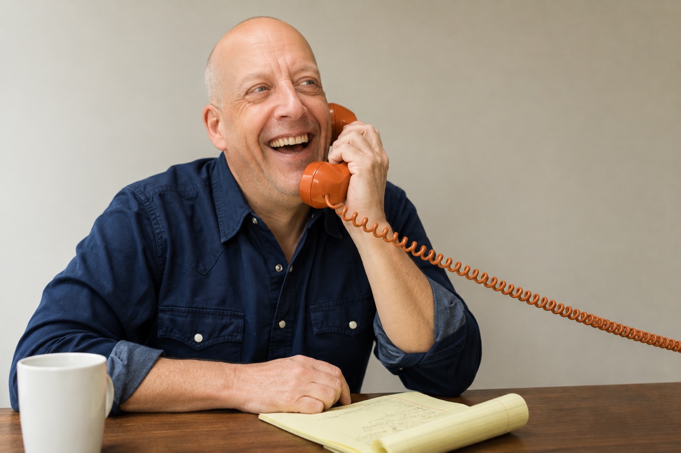 Aaron Phillips laughing on the phone at his desk, notepad and coffee in front of him